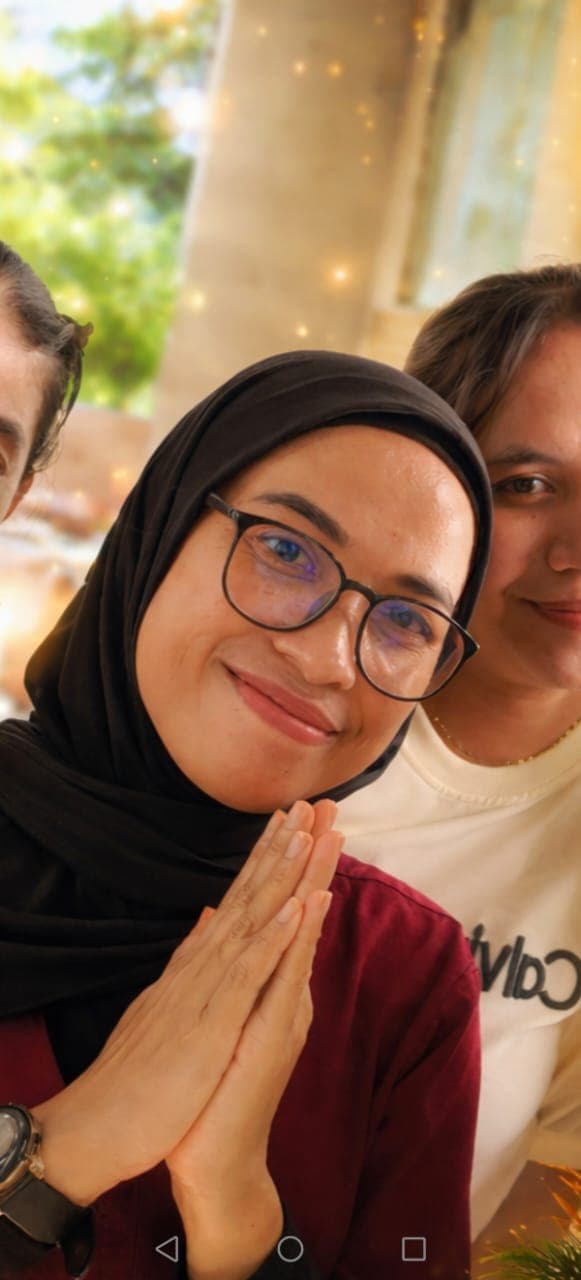 Smiling woman in a black hijab and glasses, hands pressed together in a respectful greeting.