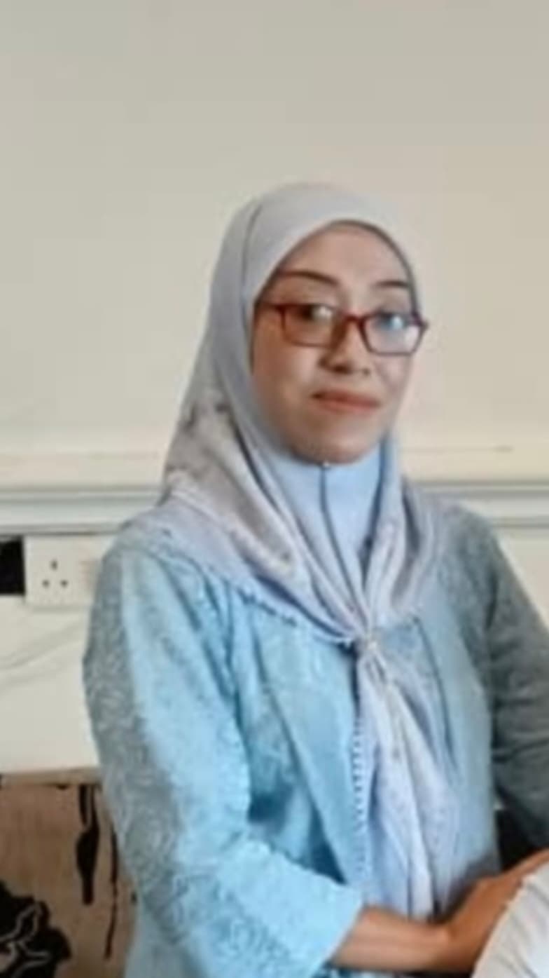 Woman wearing a light blue hijab and red-framed glasses, smiling against a plain wall.