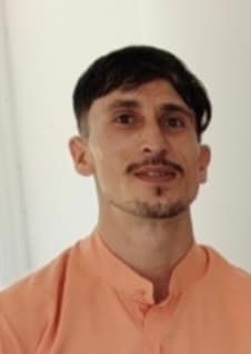 Man with dark hair and mustache wearing a peach shirt against a white wall.
