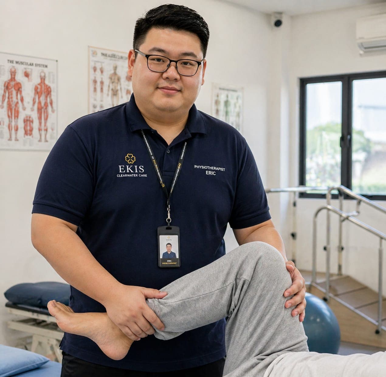 Physiotherapist Eric performs a leg stretch on a patient in a clinic with anatomical posters.